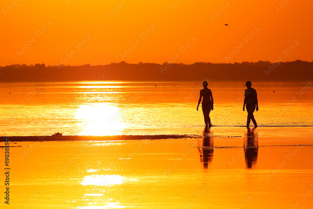 Silhouettes of two wooman (mothers and daughters) walking through the shallows of a lake against the background of a red-orange sunset