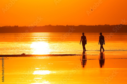 Silhouettes of two wooman (mothers and daughters) walking through the shallows of a lake against the background of a red-orange sunset