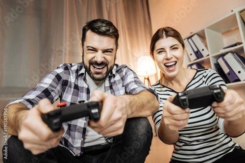 Lets play. Smiling male person sitting near his friend and leaning arms on knees while keeping console in both hands