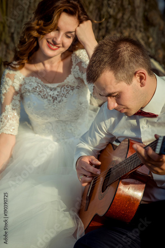 The beautiful bride with charming smile is enjoying the guitar play of the handsome groom. Outdoor wedding portrait. photo