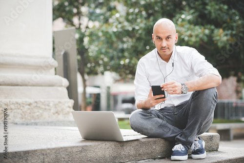 Young contemporary businessman remote working sitting outdoor in the city using smart phone and computer - portability, small business, networking concept photo