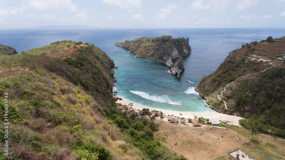 Beach with with azure water, rocky mountains and clear water at sunny day. Clear blue ocean waves rolling to the beach. Nusa Penida, Bali, Indonesia. Travel concept.