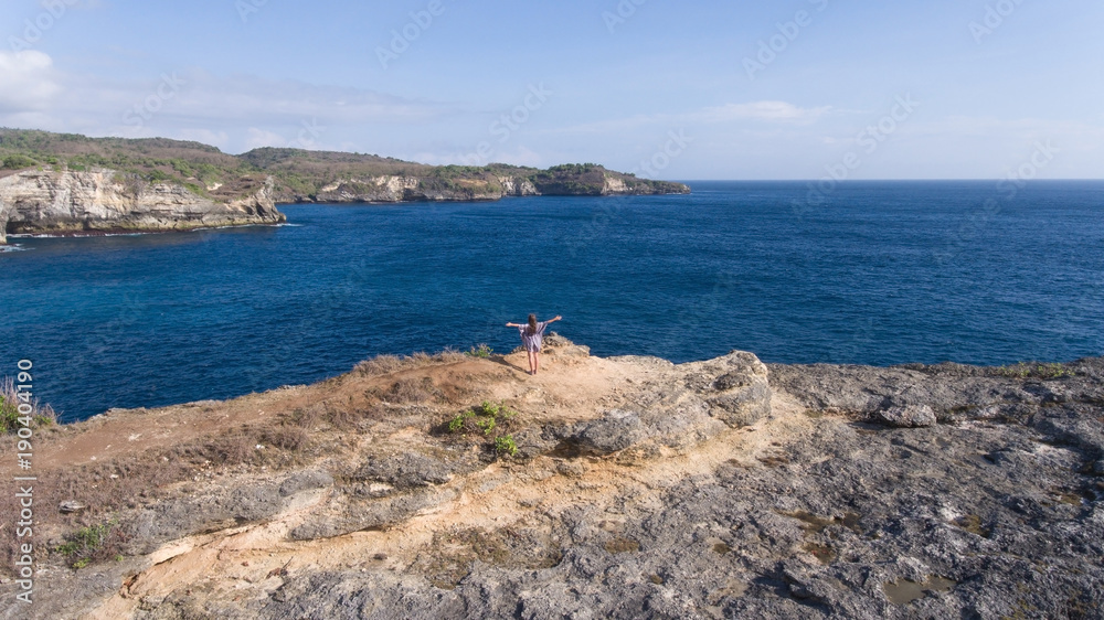 Aerial view of young girl stands on the edge of a cliff and looks at the sea Nusa Penida. Girl raising her hands up on the edge of the cliff enjoys the view of the ocean. Travel concept. Aerial view of young girl stands on the edge of a cliff and looks at the sea Nusa Penida. Girl raising her hands up on the edge of the cliff enjoys the view of the ocean. Travel concept.