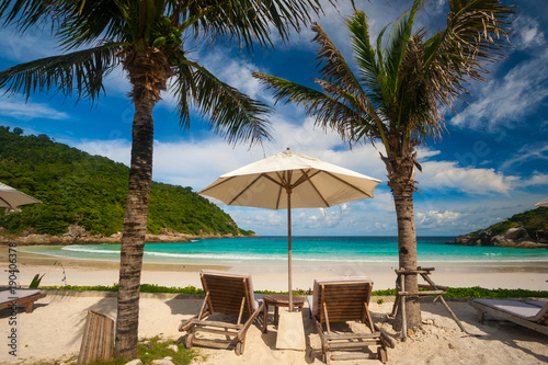 A typical beautiful holiday scene with two beach chairs and an umbrella in between, flanked by palm trees, offering a great view of Patok Beach on Racha Island, Phuket, Thailand.