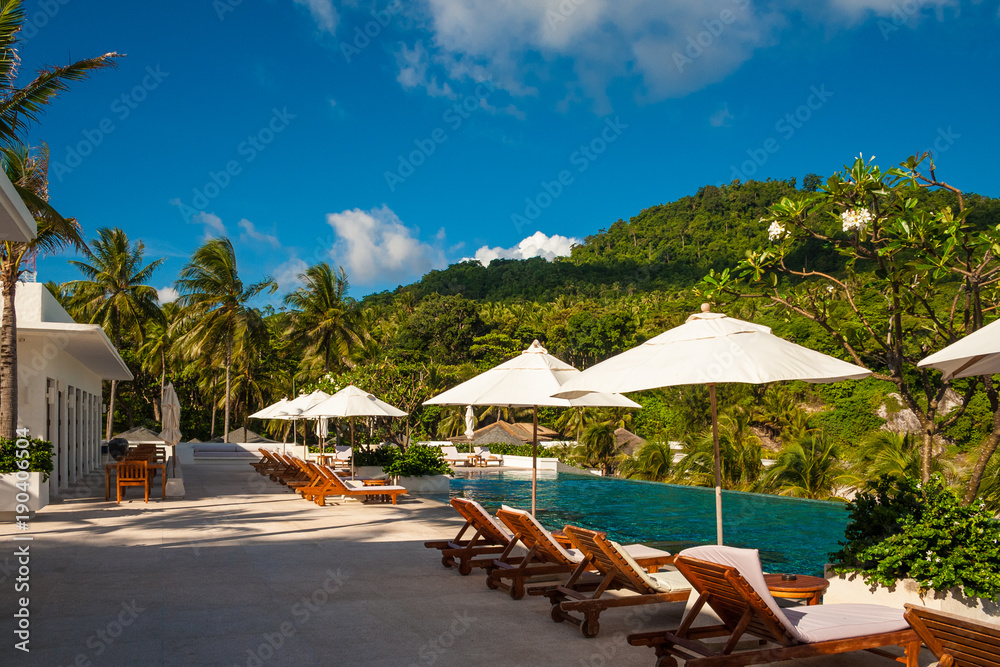 A perfect holiday photo of empty deck chairs and patio umbrellas in between, lined up in front of an infinity pool on a tropical island with a beautiful hill in the background.