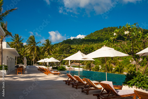 A perfect holiday photo of empty deck chairs and patio umbrellas in between  lined up in front of an infinity pool on a tropical island with a beautiful hill in the background.