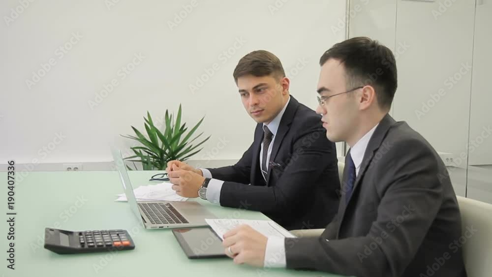 Two business people brainstorm in front of monitor at desk. In office entrepreneurs discuss project on laptop and analyze financial information. Two business people brainstorm in front of monitor at desk. In office entrepreneurs discuss project on laptop and analyze financial information.