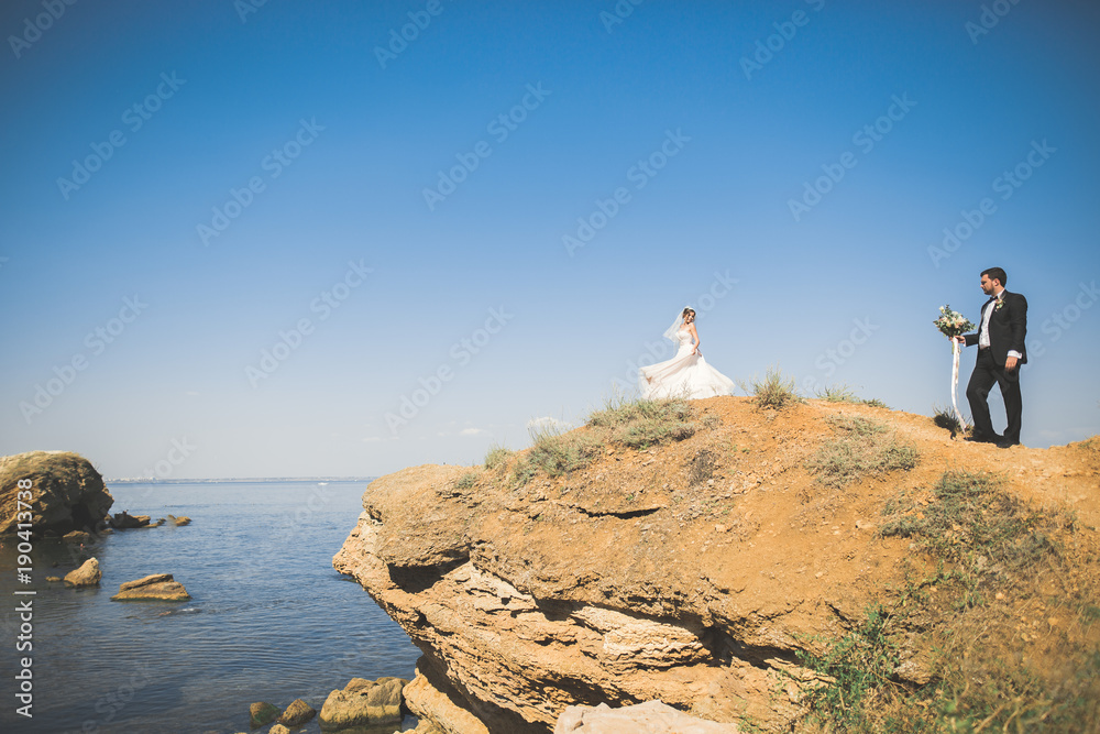 Obraz premium Wedding couple, groom, bride with bouquet posing near sea and blue sky