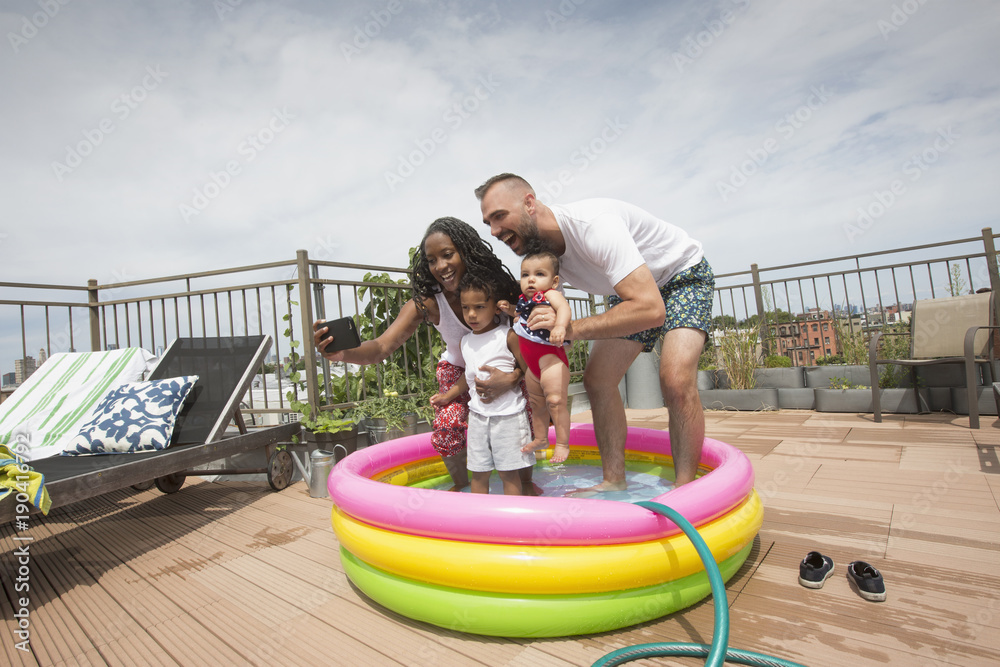 © Jasper Cole/Blend Images - Parents posing for selfie with children in inflatable swimming pool