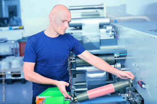 Printer operator next to the printing machine  holding printing cylinder with polymer relief plate stuck on it. Scene showing the preparation for the printing process on in-line press machine.