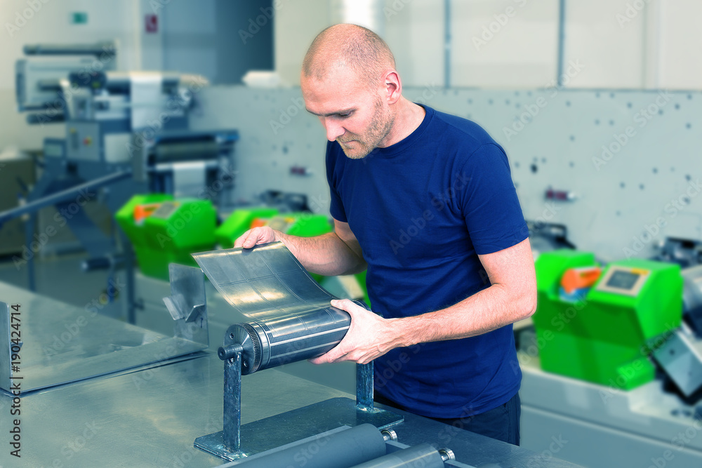 Flexo rotary die cutting preparation. Worker in printing factory, standing next to the printing machine preparing cylindrical die. Operator aligns cutting die on magnetic roll. Label manufacturing. Flexo rotary die cutting preparation. Worker in printing factory, standing next to the printing machine preparing cylindrical die. Operator aligns cutting die on magnetic roll. Label manufacturing.