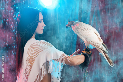 beautiful brunette girl with perfect figure standing in white shorts and transparent fashion jacket with fringe, holding the hand with bird hawk in hood on black background with smoke, fog and rain photo