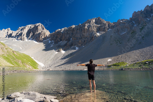 Hiker relaxing at high altitude blue lake in idyllic uncontaminated environment once covered by glaciers. Summer adventures and exploration on the Italian French Alps. Clear blue sky.