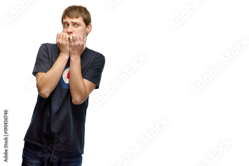 Funny guy something very frightened. A young man in a T-shirt and jeans gnaws his nails for fear. The boy made a mistake and is afraid of punishment.