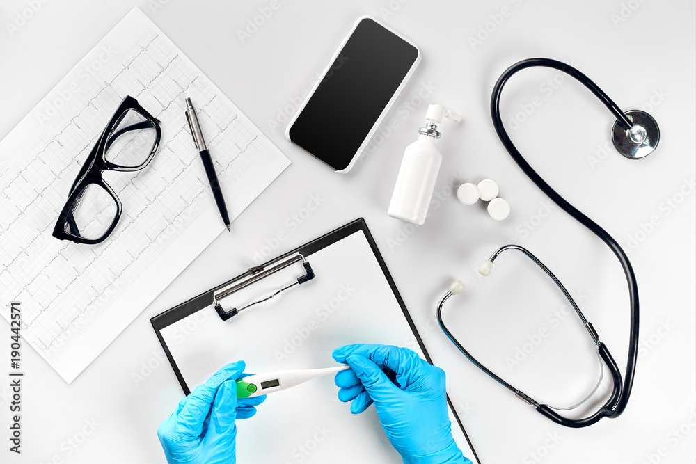 Doctor measuring temperature and holding a digital thermometer, desktop with medical equipment on white background, top view Doctor measuring temperature and holding a digital thermometer, desktop with medical equipment on white background, top view