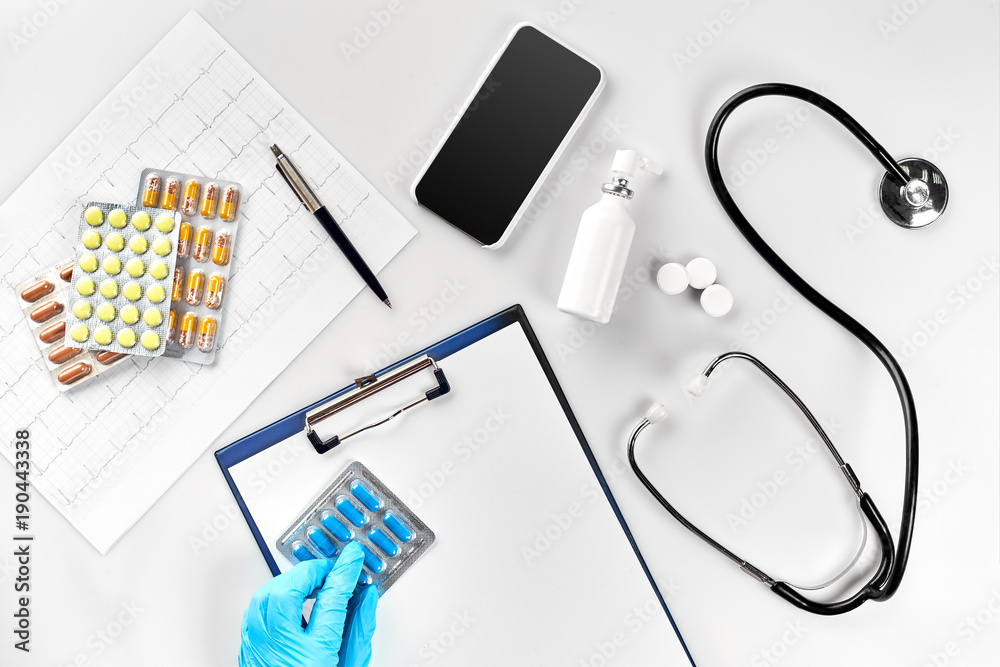 Workplace of a doctor. Pills in hand, stethoscope, clipboard and glasses on white table Workplace of a doctor. Pills in hand, stethoscope, clipboard and glasses on white table