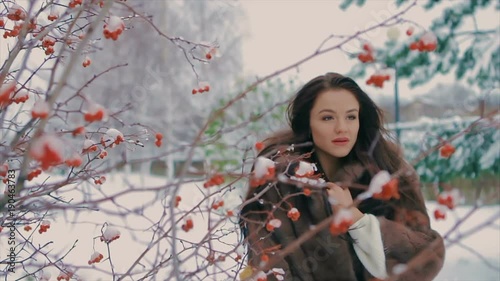 brunette stylish girl with bright makeup, near bush with red berries and Snow covered Christmas trees, in a luxurious brown fur coat walking in winter time slow motion