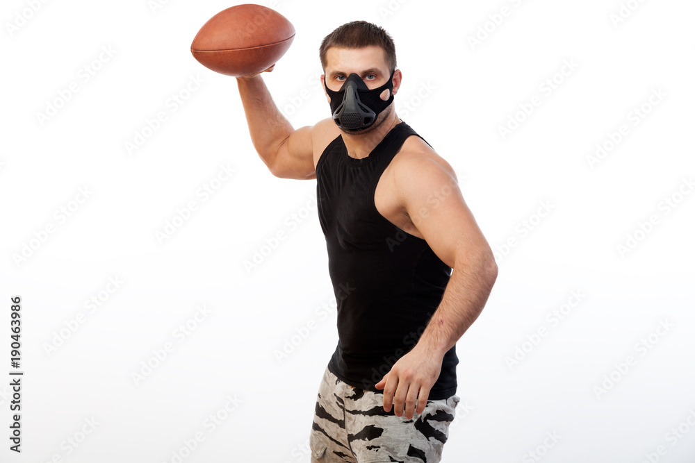 A young sporty blue-eyed man in a sporty black T-shirt and a black training mask is holding a rugby ball on a white isolated background A young sporty blue-eyed man in a sporty black T-shirt and a black training mask is holding a rugby ball on a white isolated background