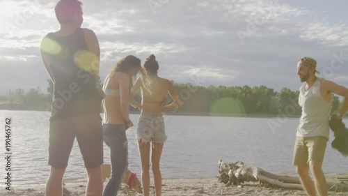 Handheld shot of happy friends undressing at riverbank against cloudy sky