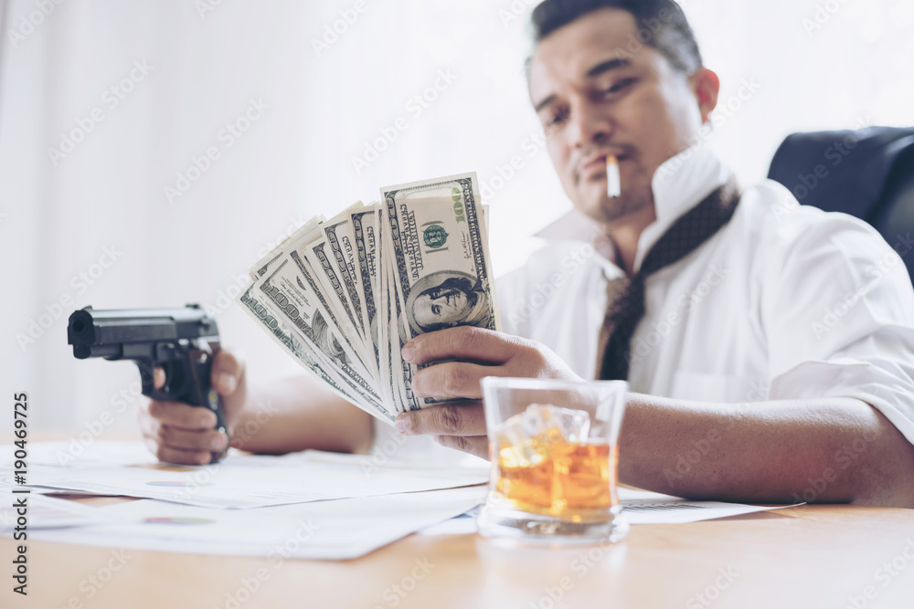 businessman at office desk holding a gun and us dollar bill businessman at office desk holding a gun and us dollar bill