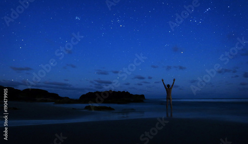 Naked man stands alone on beach at night to celebrate the clear sky of stars with hands in the air.