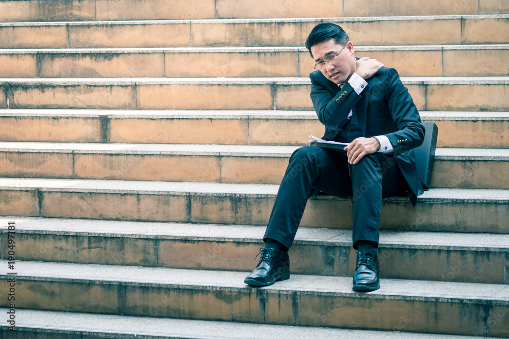 Asian business man sitting on city stairs with feeling pain in his shoulder.