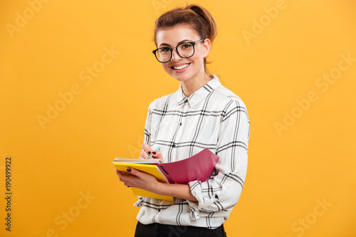 Photo of young educated woman looking on camera and writing down notes in textbook, isolated over yellow background
