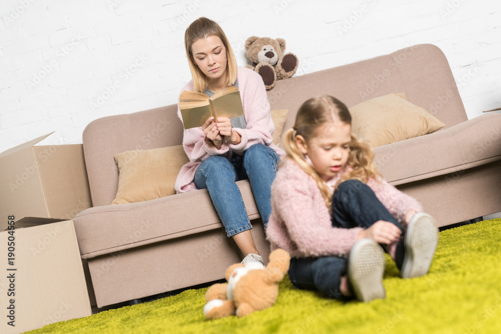 kid playing with teddy bear on carpet while mother reading book on sofa