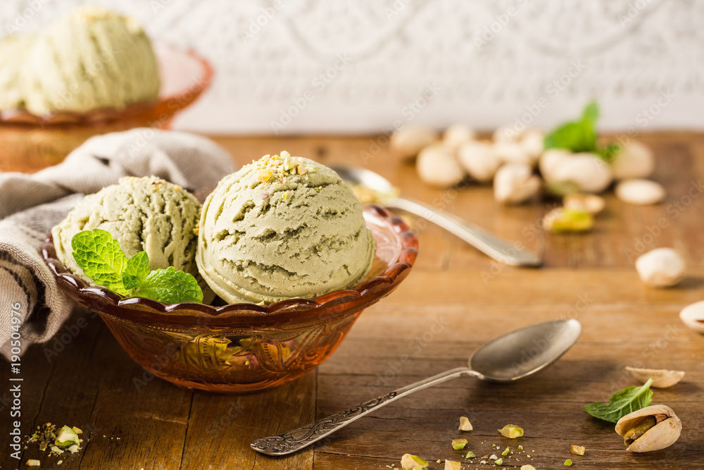 Fototapeta premium Pistachio ice cream scoops with grated pistachos in red glass bowl on old wooden table background. Homemade summer food concept with copy space.