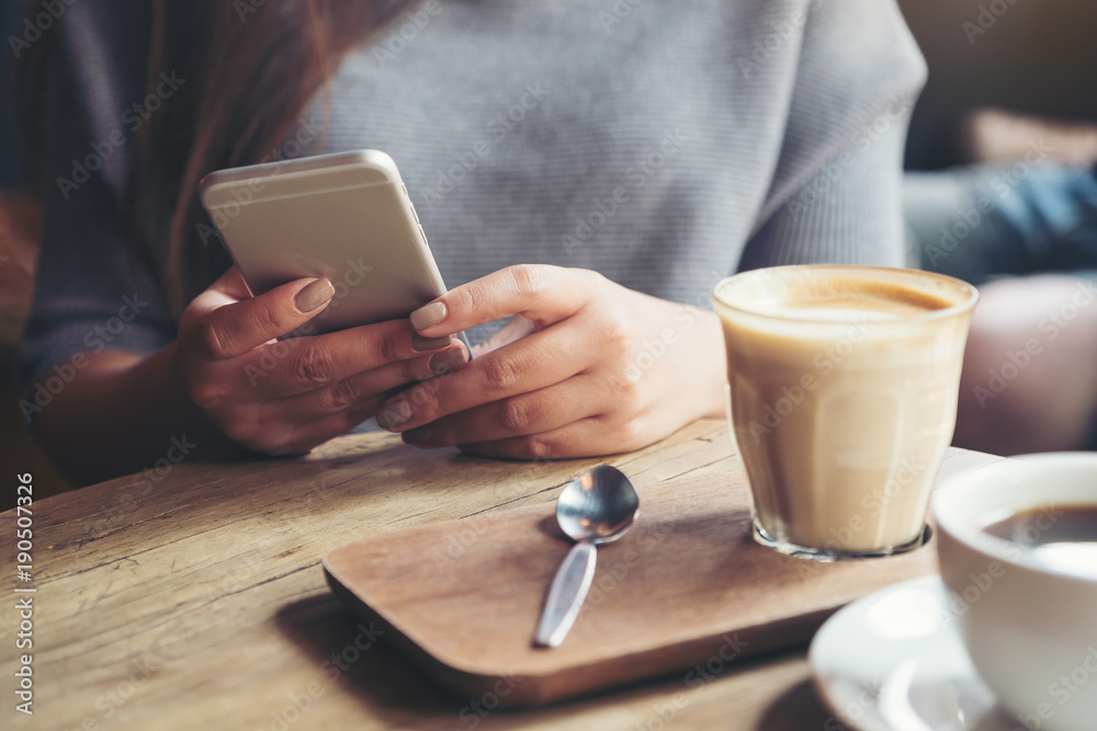 Closeup image of a woman holding and using smart phone with coffee cups on wooden table in cafe Closeup image of a woman holding and using smart phone with coffee cups on wooden table in cafe