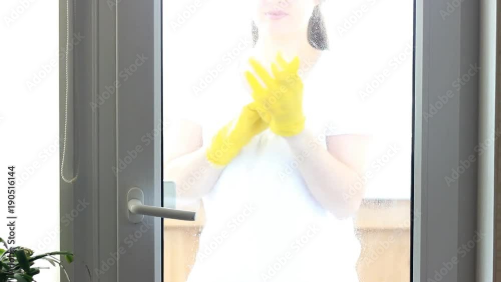 Portrait of young woman in yellow rubber gloves looks at camera smiling and washes windows and glass. The concept of cleaning, cleanliness.