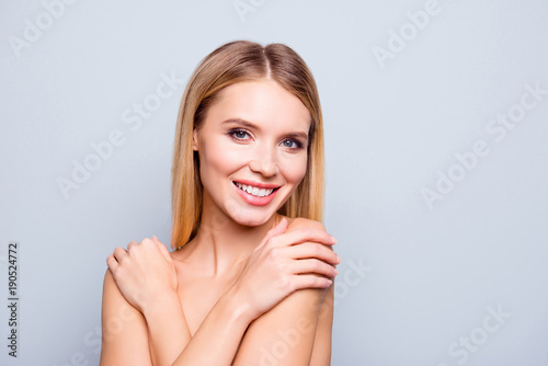 Close up portrait of healthy beautiful young woman with amazing blonde hair and clean skin. She is touching her shoulders after receiving procedure at cosmetologist. Isolated on grey background