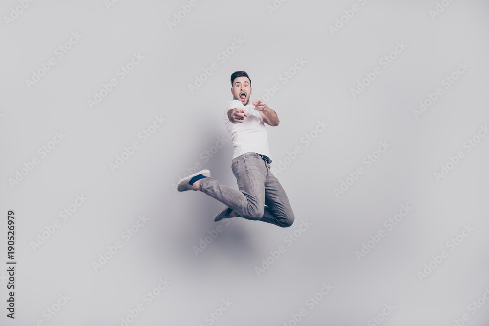 Happiness, freedom, motion and people concept - happy young bearded attractive handsome  man showing tongue with opened mouth,  jumping in air and pointing fingers to you over grey background