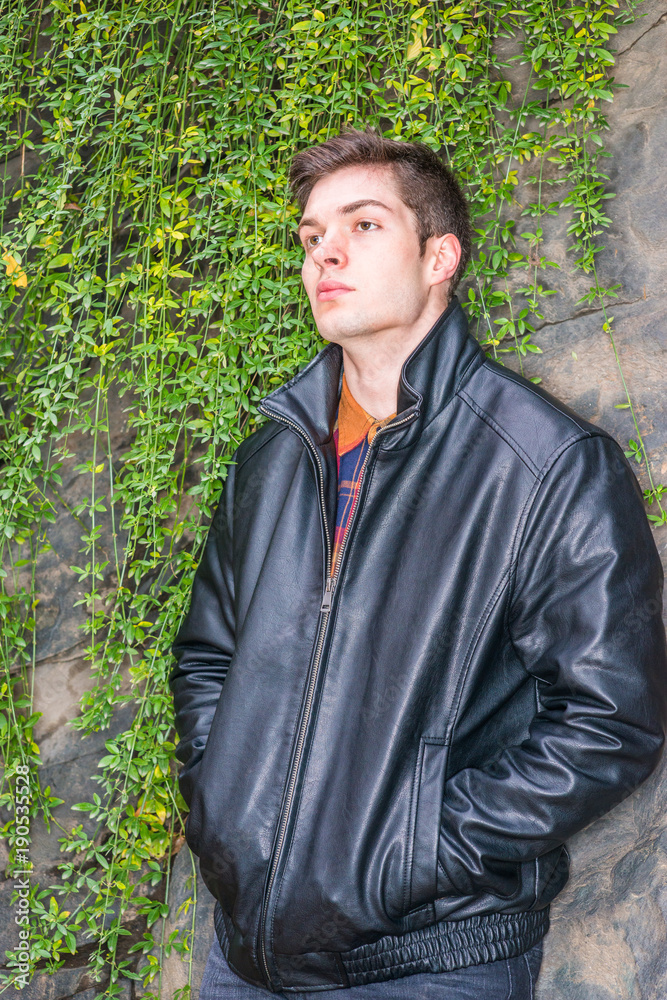 American man autumn/winter casual street fashion. Young handsome man wearing black leather jacket, standing by rock wall with long leaves at Central Park, New York, hands in pockets, thinking.. American man autumn/winter casual street fashion. Young handsome man wearing black leather jacket, standing by rock wall with long leaves at Central Park, New York, hands in pockets, thinking..
