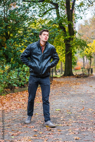 Young American man traveling at Central Park, New York in autumn day. Man wearing black leather jacket, jeans, gray casual shoes, hands in pockets, walking on road with trees, fallen leaves on ground