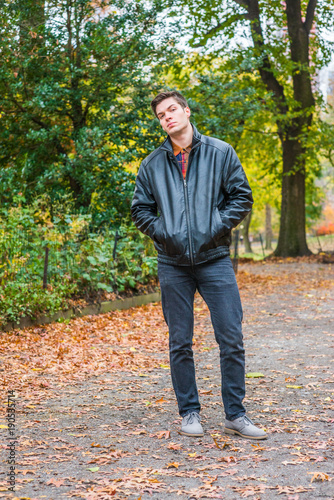 Young American man traveling at Central Park, New York in autumn day. Man wearing black leather jacket, jeans, gray casual shoes, hands in pockets, walking on road with trees, fallen leaves on ground,