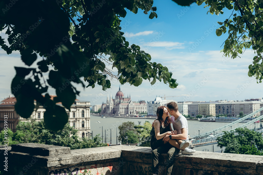 Fototapeta premium Romantic sunny portrait of the sensual couple in love tenderly rubbing noses while sitting on the balcony at the panorama view of Budapest, Hungary.