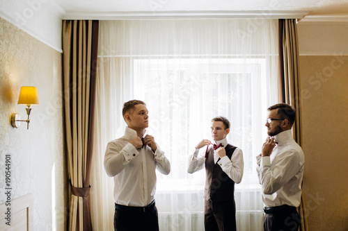 stylish groomsmen helping happy groom getting ready in the morning for wedding ceremony.
