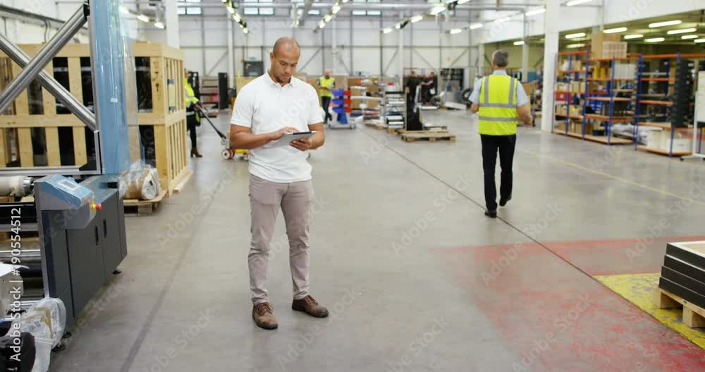 4K Young business manager in factory warehouse looking at tablet computer, with workers busy in the background