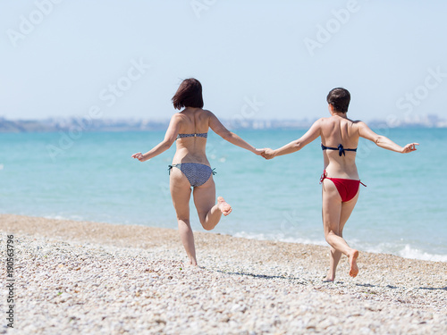 Two women in swimsuits running holding hands along beach