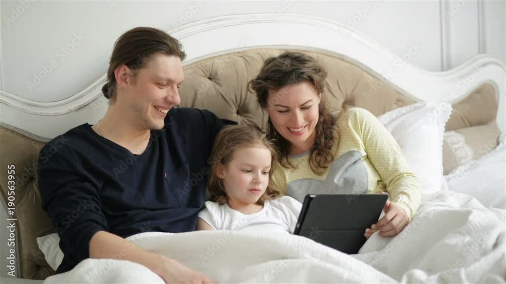 Parents and Their Daughter are Using Tablet Lying on the Bed. Young Pretty Mother with Curly Hair is Laughing. Parents and Their Daughter are Using Tablet Lying on the Bed. Young Pretty Mother with Curly Hair is Laughing.