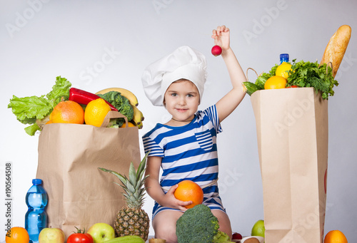 Little happy girl in the hat of the chef with big bags products holding a radish. A variety of fresh fruits and vegetables in bags on the table. photo