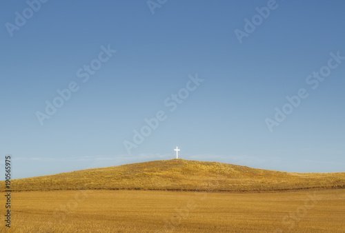 One small white wooden cross on a brown spring time hill under a blue sky in a deserted rural countryside