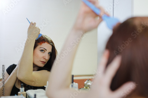 Woman in process of hair coloring at home. Applying red color to the hair and looking in the mirror.