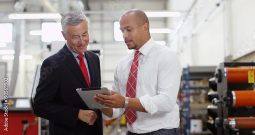 4K Businessmen greet each other & shake hands on the factory floor, looking at computer tablet & discussing operations