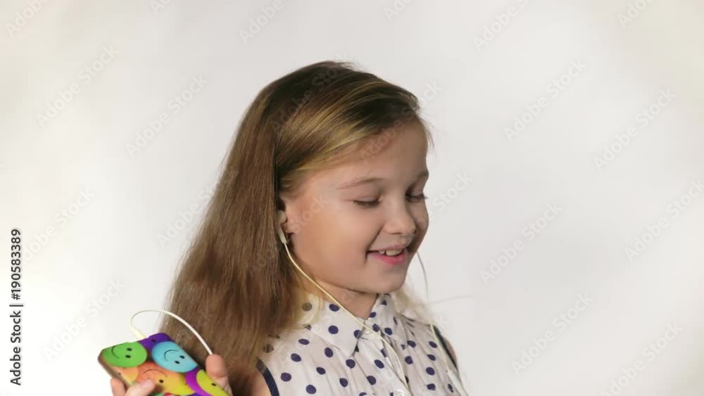 Child with earflaps dancing at studio background. The girl listens to music on the smartphone. The kid has loose long hair. Shooted on a gray white background. Child with earflaps dancing at studio background. The girl listens to music on the smartphone. The kid has loose long hair. Shooted on a gray white background.