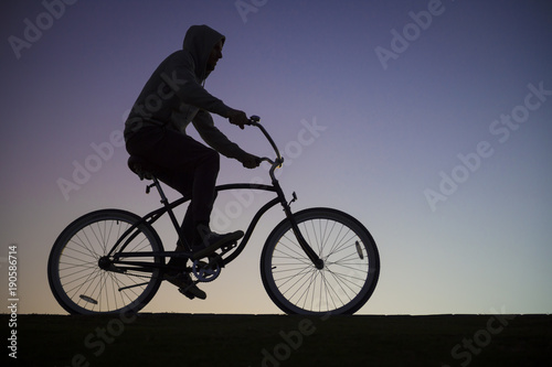 Silhouette of a man in a hoodie riding a cruiser bike in front of a purple sunset sky