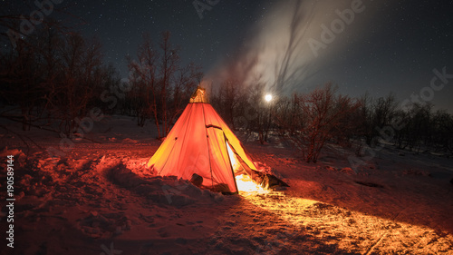 A night photo of arctic landscape with a tee-pee like tent illuminated by a fire inside projecting light out on the snow, Starry clear sky with half moon in the background. (Long exposure time)