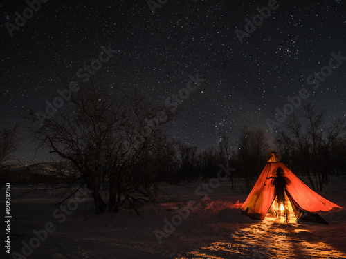 A night photo of arctic landscape with a tent and a silhouette of a person illuminated by a fire in foreground and starry clear sky in the background. (Long exposure time)