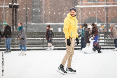 Full length portrait of caucasian joyful bearded man in yellow jacket, beige trousers, black hat on ice rink, outdoors in snowy winter day/ Weekend activities outdoor in cold weather